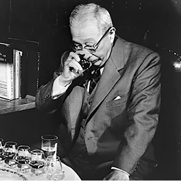 Archive photograph of Suntory founder Shinjiro Torri lifting a glass of whisky from a selction in front of him to take in its aroma