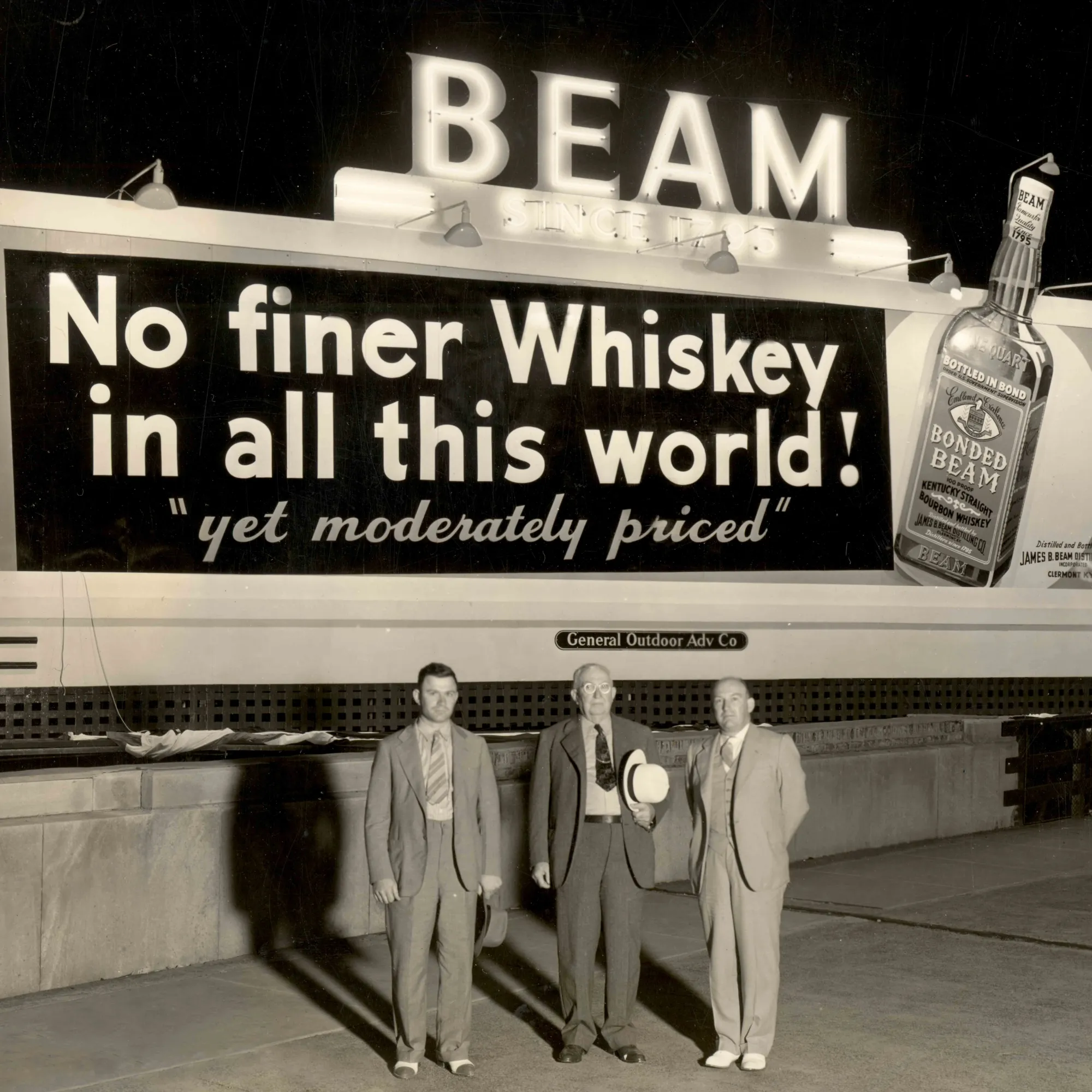 Three smartly dressed men stand before a Jim Beam billboard from the 1950s. The billboard reads: No finer whiskey in all this world. And moderately priced.