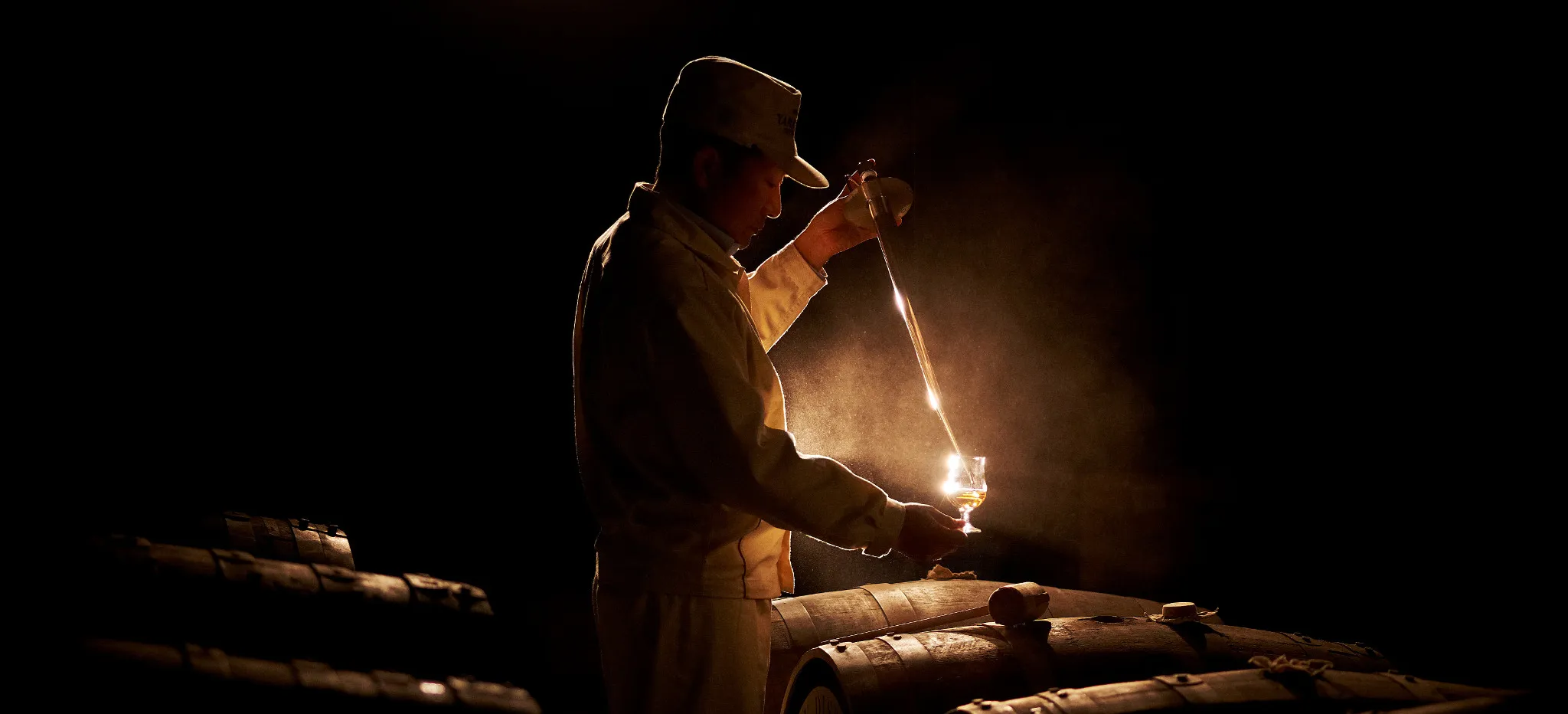 A Yamazaki distillery worker stands in a cellar surrounded by barrels. He expertly examines a glass of whisky.
