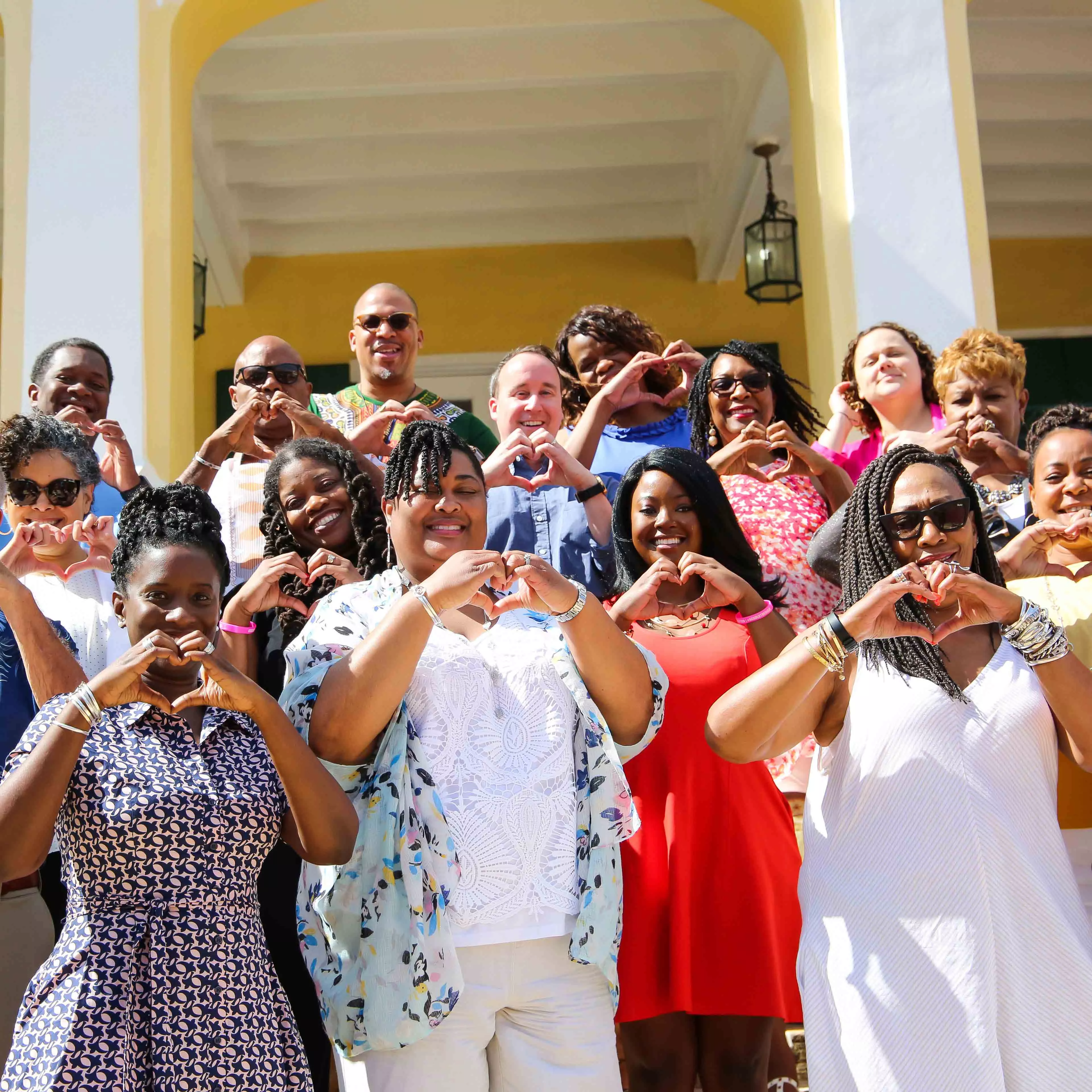 A group of smiling people stand outside on the steps on a building, holding up their hands in the shape of a heart