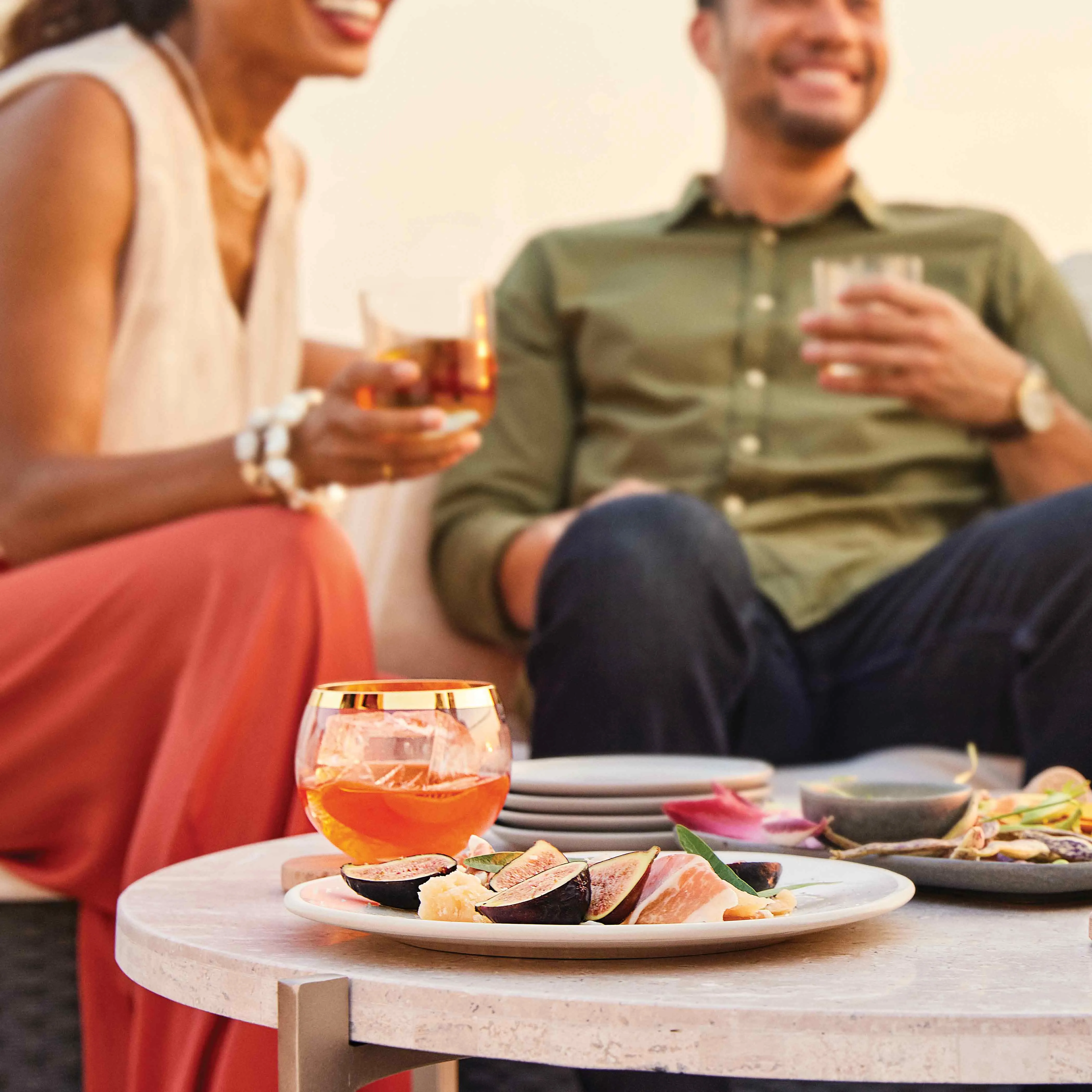 A plate of cut figs beside a small glass of cocktail. Two seated in the people hold glasses, smiling