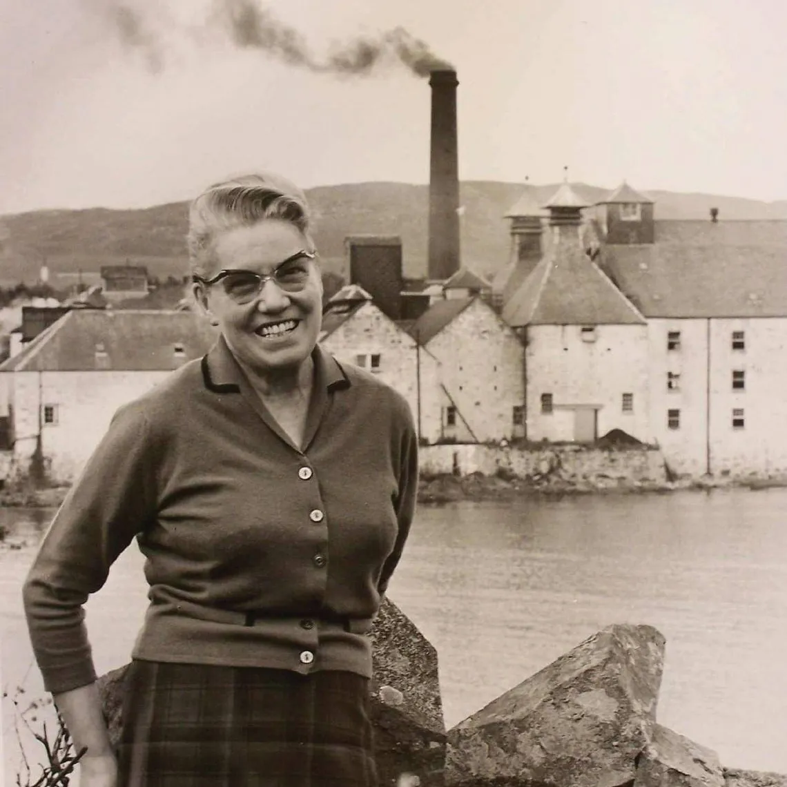 A black and white scene of a woman standing in front of a large body of water. In the background is the Laphroaig Distillery.